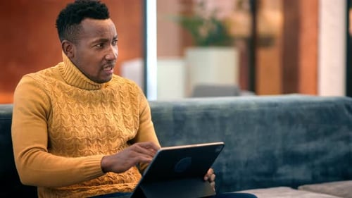 Black man working using a tablet while talking with other people, sitting on a sofa in an office