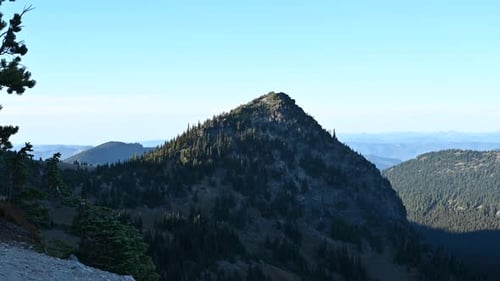 Aerial view of sunlit mountain peak surrounded by dense forests and distant ridges under a clear blu