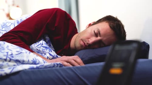 Caucasian young man in red pajamas turn off alarm clock ringing in the morning an wake up from comfo