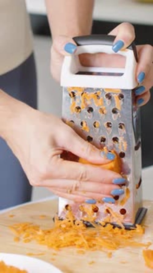 Vertical video. Woman Grating Carrot on Box Grater with Pile of Shredded Carrot Beside on Wooden