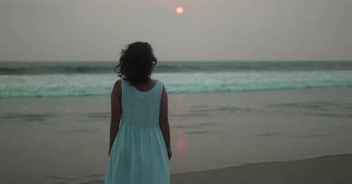 Young girl in white dress standing on beach at sunset, watching waves