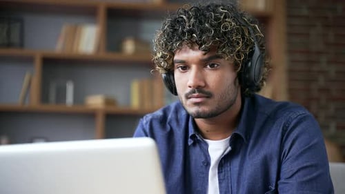 Focused IT specialist in wireless headphones works remotely on laptop sitting in living room at home