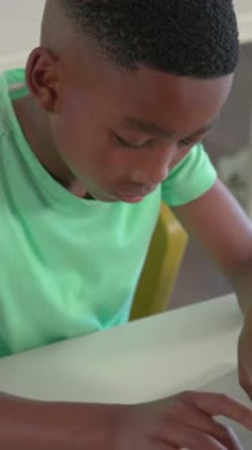 Vertical video: Zooming camera showing boy in green tee typing on laptop pausing studying in class
