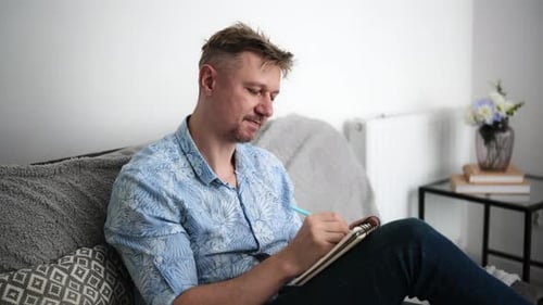 Man Writing Thoughtfully in Notebook While Relaxing on Sofa