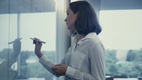 Young Female Office Worker in White Shirt is Writing on Glass Whiteboard.