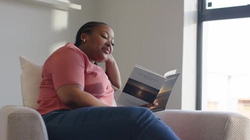 Grasping travel brochure woman flipping pages in chair at home by window while planning trip