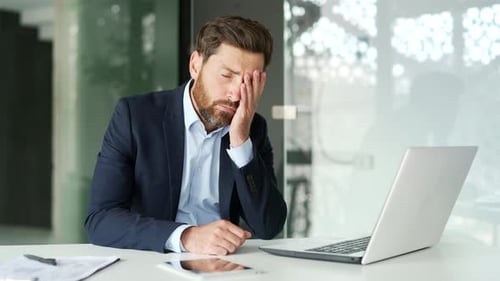 Upset businessman has depression suffering from mental pain sitting at workplace in modern office.