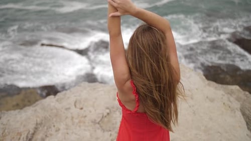 Sexy Girl In Cocktail Red Dress Standing On The Rocks And Watching The Sea Waves With Hands On Head