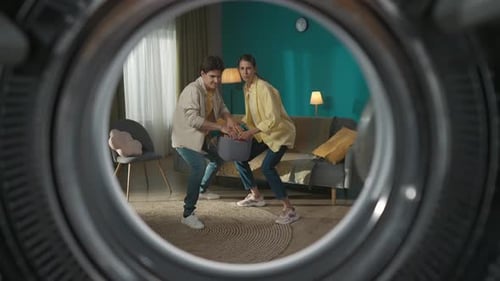 View From Inside the Empty Washing Machine Young Couple with Laundry Basket Throwing Clothes Inside