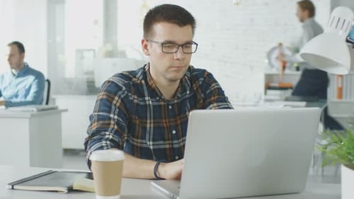 Face Front Man Sitting at His Creative Office Table Working on His Laptop. In the Background man Wo