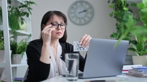 Mature Woman Drinking Pill While Sitting at Her Desk in Home Office