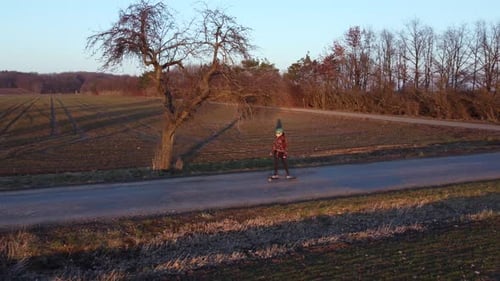 girl riding an electric longboard on a road surrounded by trees, lit by sunset in spring, aerial