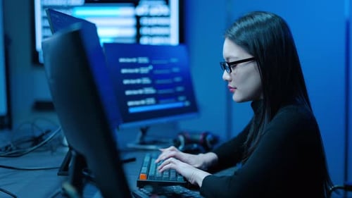 Focused Asian female programmer typing code on multiple monitors in a dark blue high-tech room.