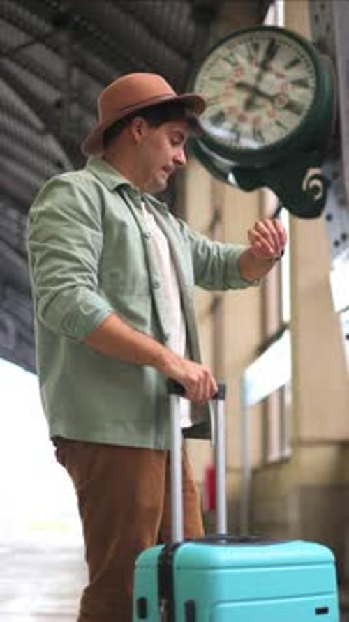vertical impatient traveler checking wristwatch while waiting by station clock for delayed train