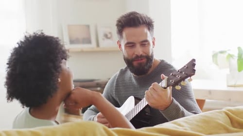 Video of rear view of happy diverse couple at home, man playing guitar, singing to woman, copy space