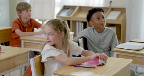 Multicultural children sit at wooden desks with notebooks and pencils, smiling during peer