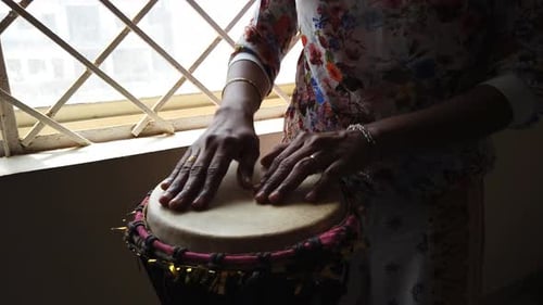 Medium wide looking down isolated shot of a woman playing djembe drum by the side of a window