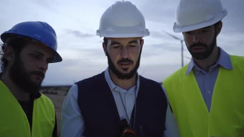 Group of Engineers Arguing Against the Turbines of a Wind Farm at Dawn Using an Electronic Tablet