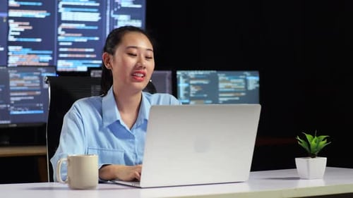Asiatische Programmiererin bei einem Meeting an einem Laptop, während sie Code auf mehreren Monitoren schreibt