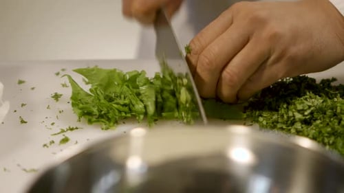 Close-up of hands finely chopping fresh herbs on a white cutting board, kitchen setting