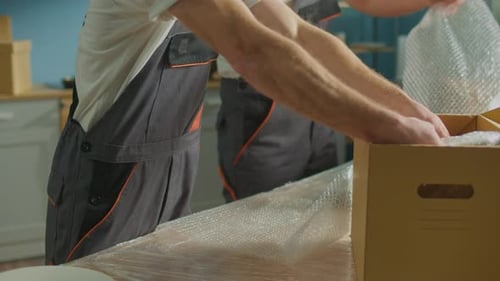 Hands Workers of Moving Company With Uniforms Carefully Packing Dishes at Boxes