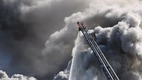 Uma mangueira de incêndio em uma escada estendida pulveriza água no fogo com uma nuvem de fumaça ao fundo