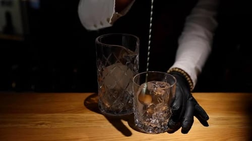 Bartender's Hands Mixing Ice in Glass Using a Mixing Spoon with a Large Ice Cube on a Wooden Bar Tab
