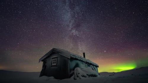 Small cabin under the milky way and aurora borealis. Cold winter night timelapse.