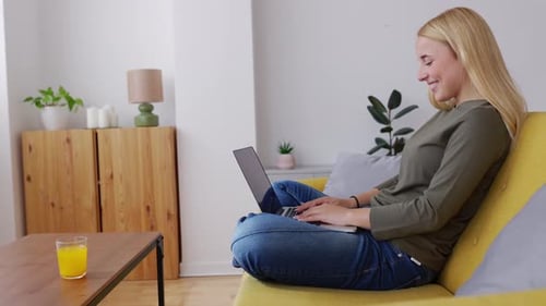 Happy Young Caucasian Woman Sitting on Sofa Working on Laptop at Home