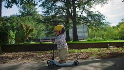 Side view of toddler riding scooter in park with yellow helmet, small child gliding forward with