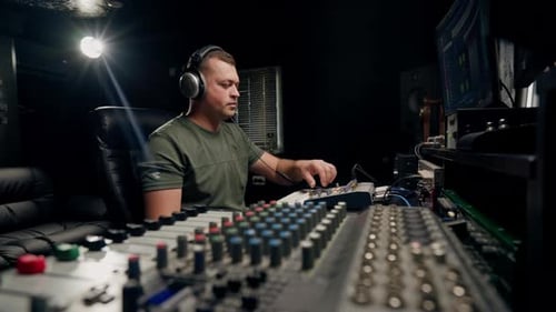 a focused male sound engineer wearing headphones sits at a mixing desk recording new track