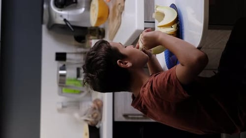 Assis à une table de cuisine, un garçon vêtu d'une chemise orange déguste une tranche de melon. Cette scène met en valeur le