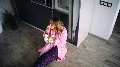 Pretty Woman Sitting on Floor in Kitchen and Eating Sweets. Satisfied Girl