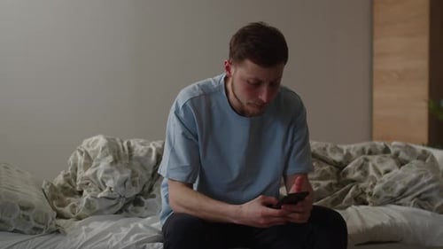 Un joven con una camiseta azul usa un teléfono inteligente mientras está sentado en la cama Un hombre hojea una red social