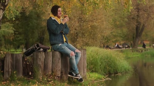 Young Handsome Man with Dreadlocks in Warm Clothes Drinking Coffee Near Lake in Golden Autumn