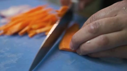 Slicing peeled carrots into strips with a large kitchen knife on a blue cutting board, close-up.