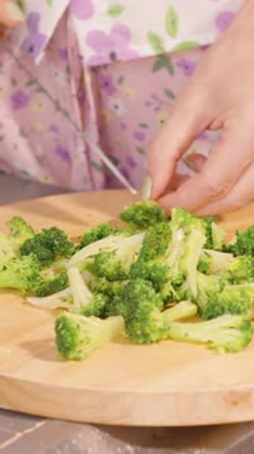 Vertical video. Woman in Floral Apron Chopping Fresh Broccoli on Round Wooden Board at Kitchen