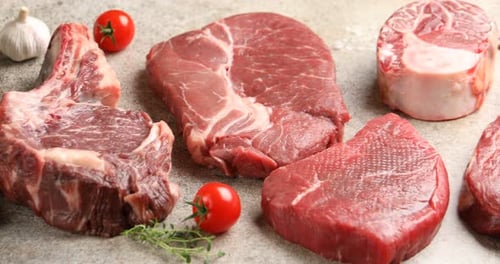 Stücke von rohem Rindfleisch, Gewürzen und Tomaten auf grauem Tisch, Nahaufnahme. Kamera bewegt sich nach rechts