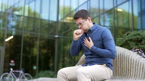 Sick businessman coughing sitting on a bench on the street near office building. Upset bearded man