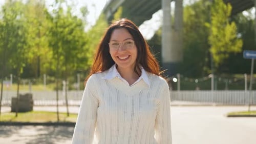 A Beautiful Young Woman with Glasses Smiles Looks at the Camera and Walks Outside on a Sunny Day
