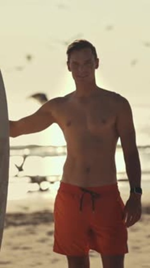 Surfer Holding Surfboard on Beach at Sunset with Seagulls