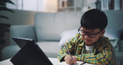 Asian boy wearing glasses schoolboy learning online with a digital tablet and writing on worksheets