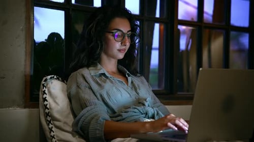 Young Woman in Home Office Looks at Laptop Screen and Works Typing on Keyboard