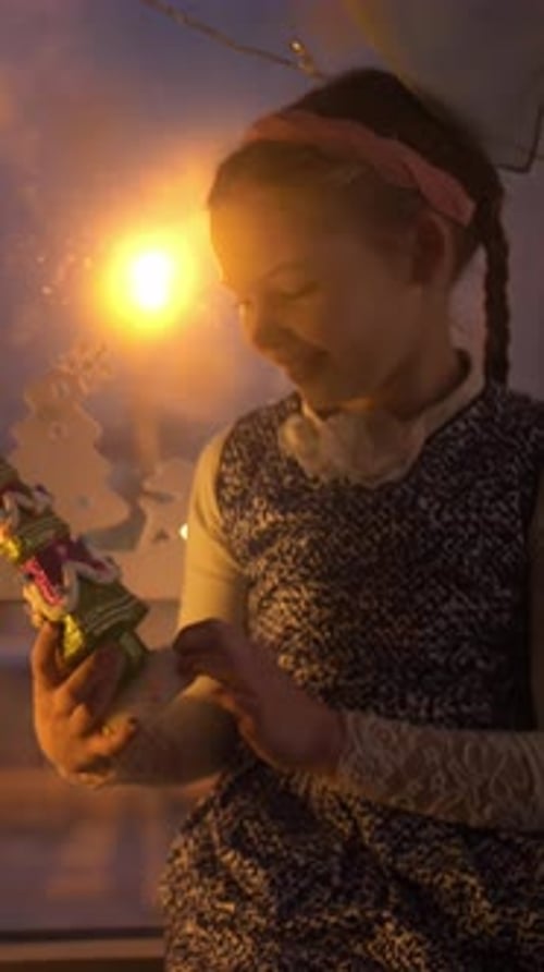 Jeune fille tenant un ornement festif tout en regardant par la fenêtre éclairée par une lumière chaude créant un