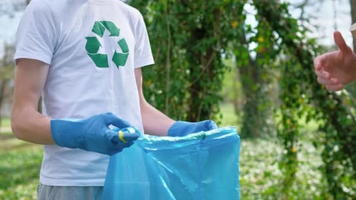 Two men collecting plastic garbage in bag in a polluted park. Rubber gloves, recycling sign on T-shi