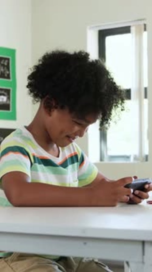 Vertical video: Playing student in striped tee tapping handheld device at classroom desk, smiling