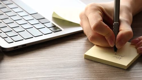 Mujer escribiendo una nueva contraseña en una nota adhesiva en un escritorio de madera, primer plano