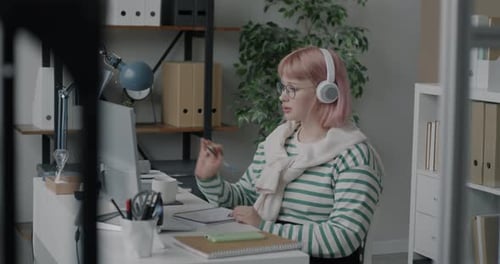 Young Woman Making Online Video Call with Computer and Headphones Talking Writing at Desk in Office