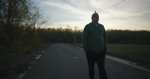 Man walking on an empty rural road at sunset, hands in pockets, enjoying peaceful evening in a
