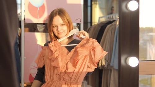 Mulher alegre navegando em um vestido rosa no cabide inspecionando roupas perto de um espelho iluminado no varejo